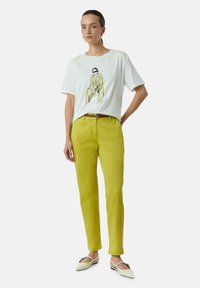 Witte T-shirt met een geïllustreerd ontwerp, gecombineerd met felgele broek en witte schoenen met een decoratieve sluiting.