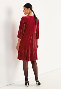 Samtrotes Kleid mit Dreiviertelärmeln und einem gestuften, rüschierten Saum. Glatte Textur und eng anliegendes Oberteil, kombiniert mit schwarzen Absätzen und Strumpfhosen.
