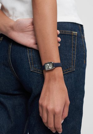 Person wearing blue jeans and white top with hand resting on lower back, showing a square-faced silver wristwatch with navy strap.