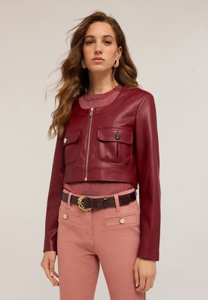 Veste en cuir bordeaux raccourcie avec deux poches avant et une fermeture éclair. Portée par-dessus un haut rose ajusté et associée à un pantalon rose et une ceinture marron.