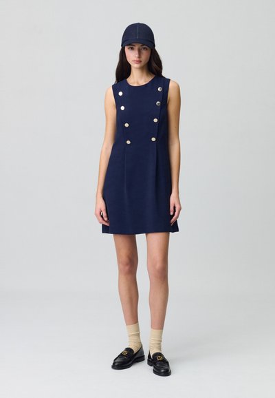 Vestido sin mangas en color navy con botones dorados, silueta recta y textura suave. Combinado con mocasines negros y calcetines beiges.