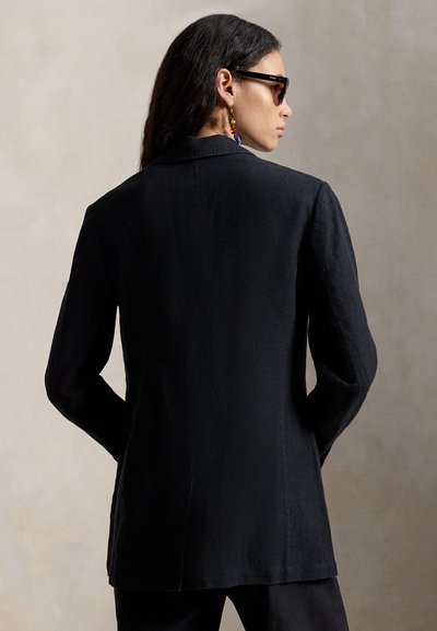 Osoba s dugom tamnom kosom u crnom sako, crnim hlačama, sunčanim naočalama i šarenim visećim naušnicama, promatrana s leđa na jednostavnoj pozadini.