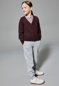 Jong meisje dat glimlacht, met handen in haar zakken, draagt een donkerpaarse cardigan, gestreept shirt, grijze trainingsbroek en witte sneakers met klittenbandsluitingen.