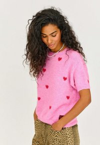 Ružový sveter s krátkym rukávom a červenými srdcovými akcentmi, vyrobený z mäkkého materiálu. Skombinovaný s nohavicami s leopardím vzorom, ktoré majú bočné vrecko.