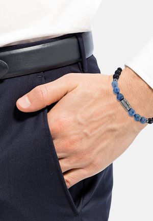 Pulsera de cuentas con piedras azules y negras y un acento metálico, llevada en una mano que descansa en un bolsillo de pantalón azul marino, junto a un cinturón negro.