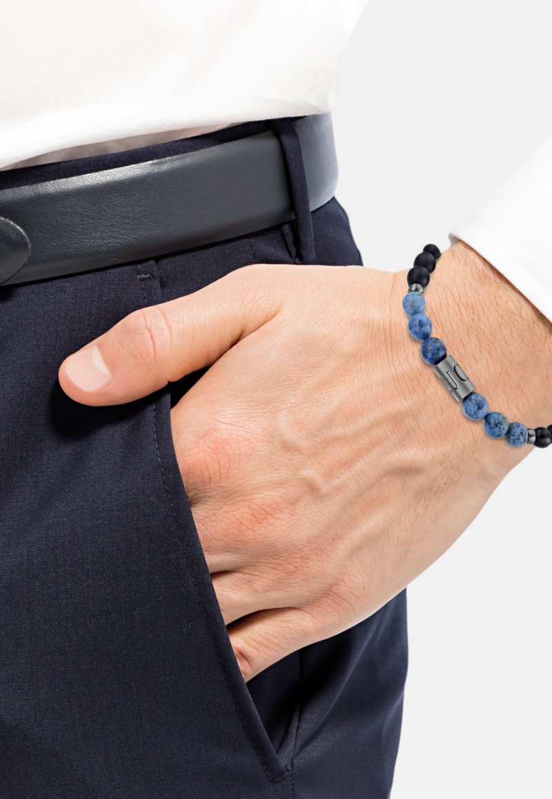 Braccialetto di perle con pietre blu e nere e un accento metallico, indossato su una mano che riposa in una tasca di pantaloni blu navy, accanto a una cintura nera.