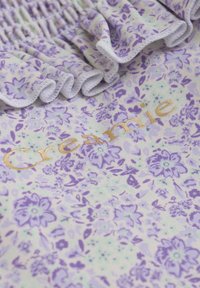 Blommig klänning med ett lavendelmönster på ljus bakgrund, med fransade kanter och guldtext som säger "Creamie."