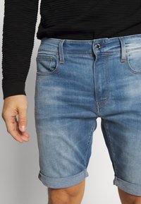 Vaaleansiniset farkkushortsit, joissa on käännetyt lahkeet, viisipokkaiset taskut, metallinen nappisuljenta ja hienovarainen kulutuskäsittely kankaassa.