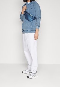 Denimblå hoodie med justerbar huva, vita oversized byxor och grå New Balance-sneakers. Har en glänsande blå axelväska.