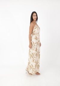 Robe maxi à bretelles spaghetti, présentant un motif floral dans des teintes beige, blanche et verte ; tissu léger, taille cintrée, design fluide.