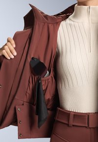 Burgundi kabát sima felülettel és patent gombokkal, egy zsebbel, amelyben egy fekete tárgy és egy fekete szalag részlet található, krémszínű bordázott felsővel párosítva.