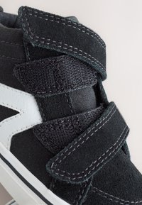 Zwarte hoge sneaker met witte zijstreep en twee brede klittenbandsluitingen op een witte achtergrond.