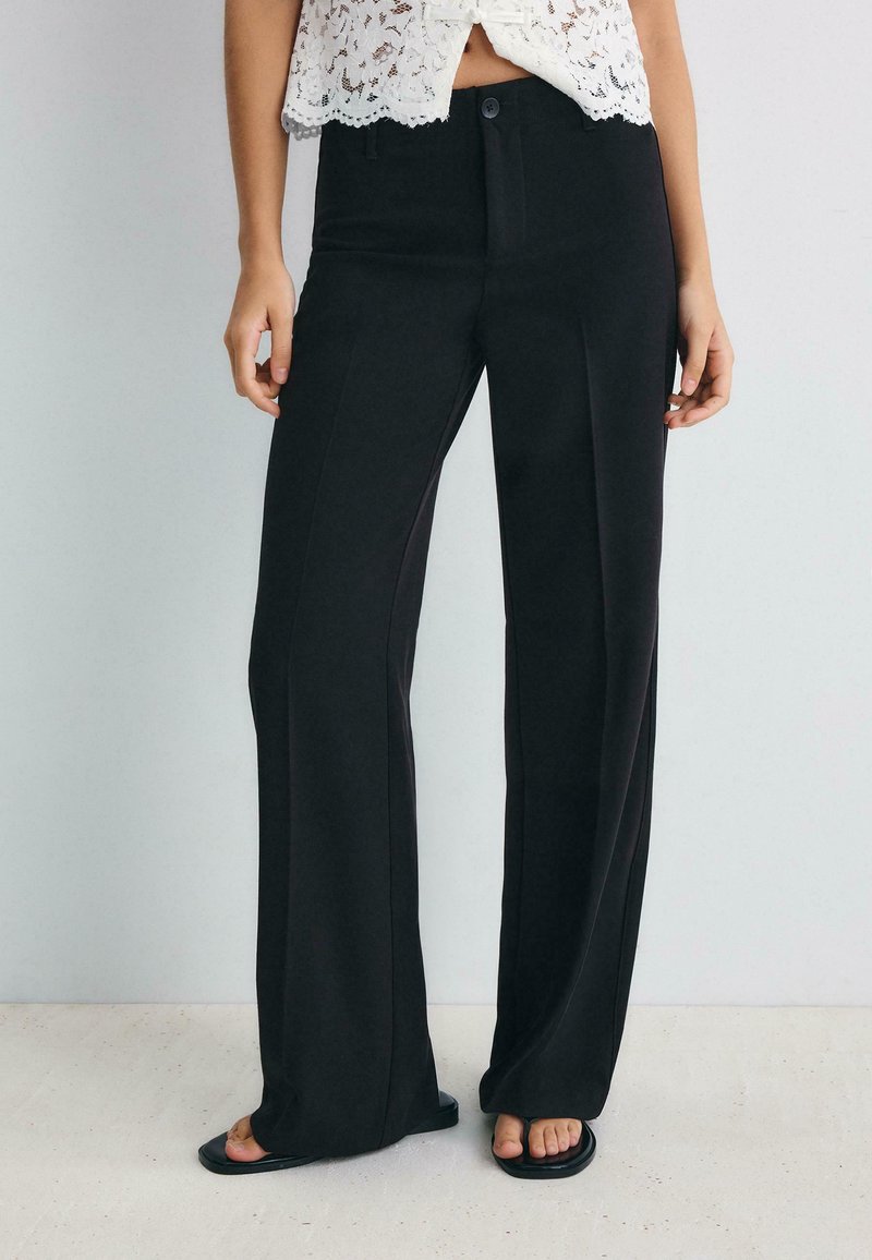 Femme portant un pantalon noir à jambes larges et des sandales noires, associé à un top court en dentelle blanche sur un fond uni.