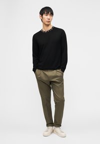 Homme portant un haut noir à manches longues avec un col coloré, pantalon plissé vert olive, baskets blanches, debout les mains dans les poches.