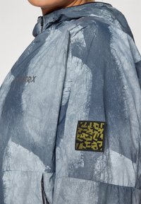 Wasserabweisende Jacke in Blau und Grau mit strukturierter Oberfläche. Verfügt über eine Kapuze und einen schwarzen Patch mit gelben geometrischen Mustern.