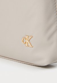 Geantă din material bej cu o textură netedă și logo-ul "CK" în ton de aur. Dispune de o formă rectangulară și detalii subtile de cusătură.