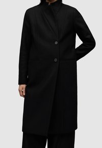 Czarny wełniany płaszcz o dopasowanym kroju, z wysokim kołnierzem i dwoma guzikami z przodu. Posiada boczne kieszenie i gładką fakturę.