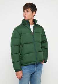 Veste matelassée verte avec fermeture éclair, col montant et texture matelassée. Comprend des poches latérales et un design ajusté, portée par-dessus une chemise grise.