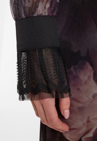 Schwarzes Spitzenarmband mit gewelltem Rand, übereinander geschichtet auf durchsichtiger Stoff. Texturierter Netzuntergrund mit floralen Mustern, die darunter sichtbar sind.