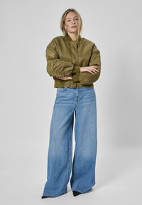 Olivgrüne Bomberjacke mit gerippten Bündchen, kombiniert mit weit geschnittenen, hellblauen Jeans. Schlichtes Design, glatte Textur und minimale Akzente.