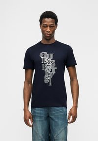 Άνδρας που φοράει μπλε μπλουζάκι Guess με κάθετο λευκό κείμενο "GUESS EST 1981" και μπλε τζιν, στέκεται μπροστά σε απλό λευκό φόντο.