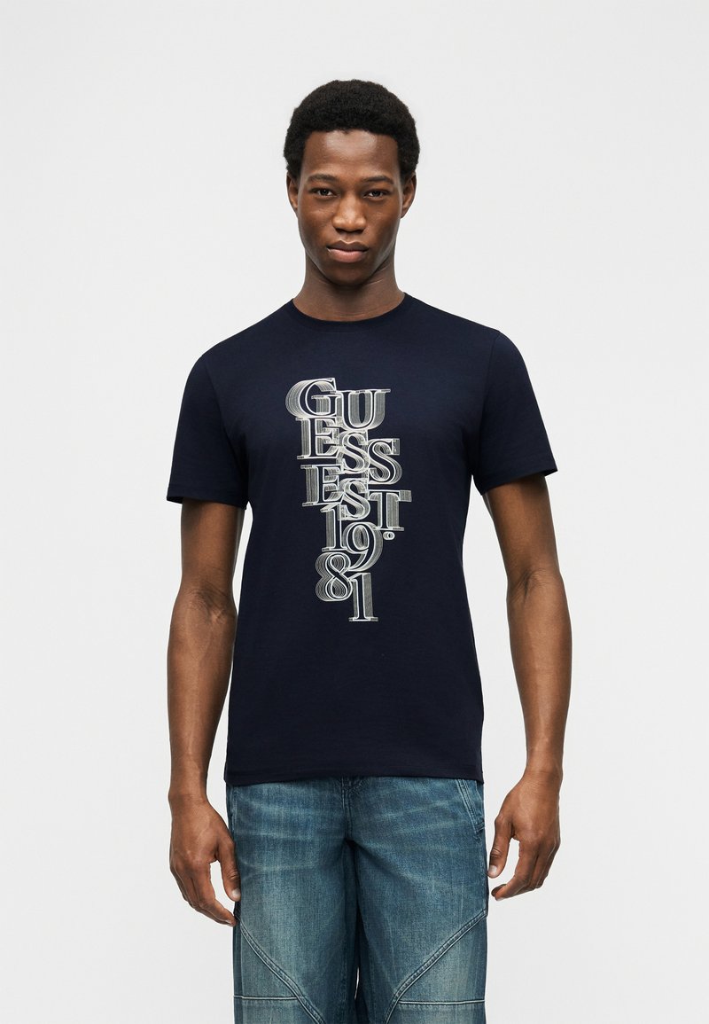 Άνδρας που φοράει μπλε μπλουζάκι Guess με κάθετο λευκό κείμενο "GUESS EST 1981" και μπλε τζιν, στέκεται μπροστά σε απλό λευκό φόντο.