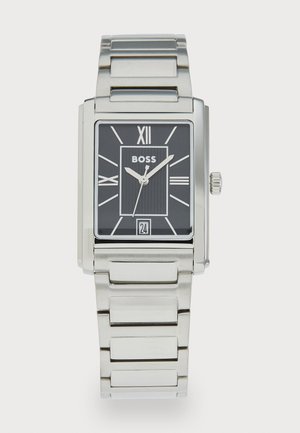 Montre rectangulaire argentée avec un cadran noir, des chiffres romains, un centre texturé et une fenêtre de date. Elle présente un bracelet en maillons métalliques.