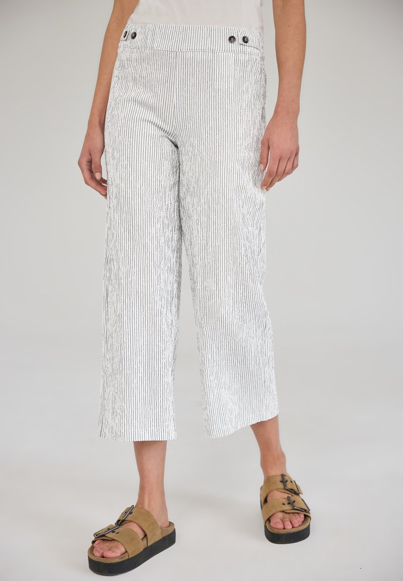 Personne portant un pantalon blanc à larges jambes rayé et raccourci, ainsi que des sandales marron à double boucle, se tenant devant un fond neutre.