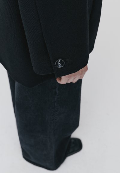 Blazer in lana nera con finitura texturizzata, caratterizzato da un bottone nero e una vestibilità leggermente oversize, abbinato a pantaloni in denim scuri a gamba larga.