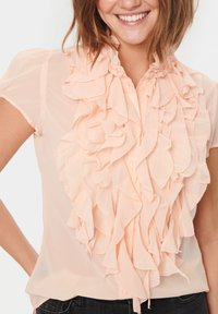 Blusa in chiffon pesca con maniche corte e un fronte arricciato. Presenta una chiusura con bottoni e una texture leggera e fluida.