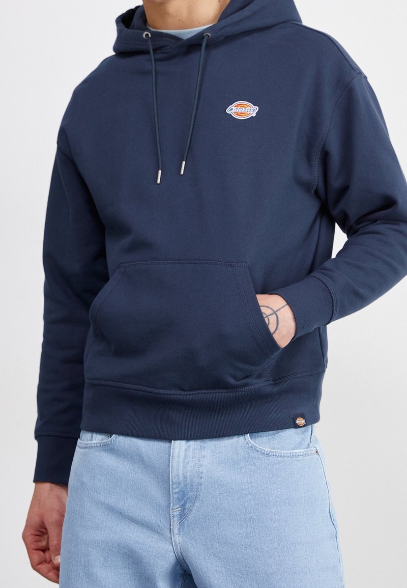 Homme portant un sweat à capuche bleu marine avec poche avant et logo orange, associé à un jean bleu clair, main partiellement dans la poche.