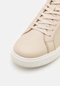 Baskets en cuir beige avec une texture lisse, semelle en caoutchouc blanc plat et lacets plats assortis, présentant des détails de couture subtils sur le côté.