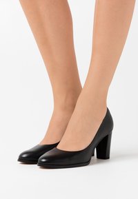 Escarpins noirs en cuir à talons hauts avec un bout arrondi, une texture lisse, et un talon épais, présentant un design simple et classique.