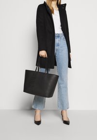 Black tote bag with embossed logo, made of smooth faux leather. Featuring dual flat handles and a structured design, paired with casual denim.