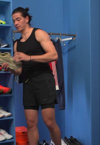 Tênis atlético cinza claro segurado por uma pessoa vestindo um top preto e calções, em pé junto a ténis coloridos em prateleiras azuis numa sala de provas.