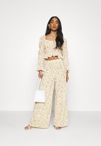 Top cropped amarelo com padrão floral, mangas com babados e calças largas a condizer. Modelo segura uma pequena bolsa branca.
