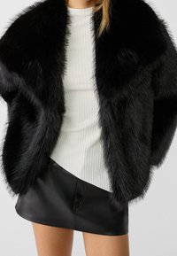 Black faux fur coat with a soft texture, featuring a wide collar and open front. Underneath, a white ribbed top and black leather mini skirt.