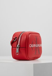 Červená kompaktní taštička Calvin Klein Jeans se stříbrným zipem a černými písmeny "CK" na přední straně, vystavená na bílém podstavci.