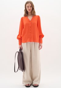 Bright orange V-neck blouse with button detail, paired with loose beige linen pants and a dark brown handbag. Flat black sandals.