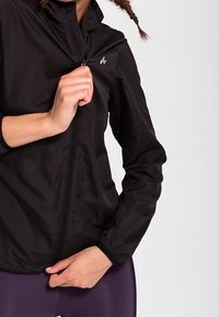 Czarna lekka kurtka z przednim suwakiem, wysokim kołnierzem i elastycznymi mankietami; z małym logo na lewej stronie klatki piersiowej.