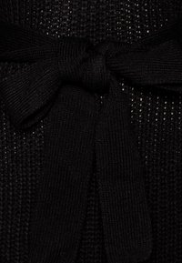 Černá pletená tkanina s texturovaným vzorem; obsahuje detaily vázaného pásku vyrobené ze stejného materiálu, což obohacuje design oděvu.