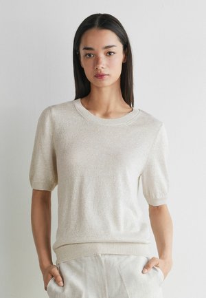 Kvinde med langt mørkt hår iført en lysebeige kortærmet striktop og matchende bukser, stående med hænderne i lommerne mod en ensfarvet væg.