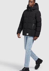 Schwarze Steppjacke mit Kapuze, Reißverschluss und gestepptem Design. Verfügt über seitliche Taschen und ein Logo-Patch am Saum. Kombiniert mit hellen Jeans und Stiefeln.