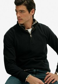 Czarna bluza z zamkiem na wysokości szyi i koszulą w kratę pod spodem. Posiada subtelne logo oraz ściągacze u rękawów. Gładka tekstura.