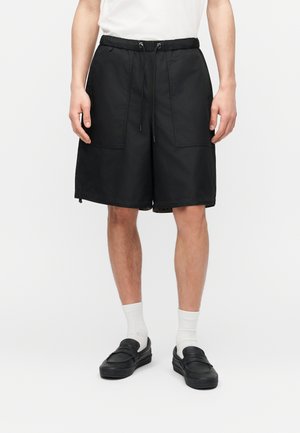 Mężczyzna ubrany w czarne szorty za kolano z przednimi kieszeniami, białe skarpetki, czarne wsuwane buty oraz białą koszulę z krótkim rękawem.