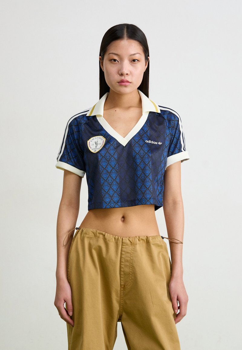 Avklippt blå adidas-tröja med diamantmönster, vit krage och logotyp-patch. Kombinerad med beige byxor med dragsko.
