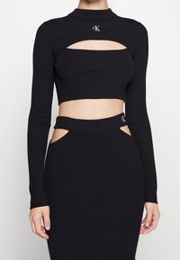Schwarzes, geripptes Strick-Crop-Top mit langen Ärmeln, das über einen Stehkragen und Cut-Out-Details verfügt. Dazu gehört ein passender Rock mit seitlichen Cut-Outs.
