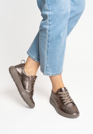 Person wearing metallic bronze lace-up sneakers with side zipper and light blue cropped jeans, posing with one foot slightly raised.