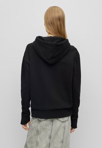 Žena s dlouhými blond vlasy oblečená v černé mikině a světle šedé džínové sukni, viděná zezadu na jednobarevném pozadí.