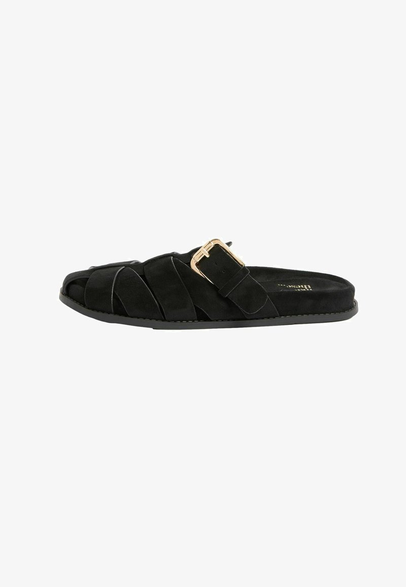 Sandalo piatto nero con cinturini incrociati in suede e fibbia dorata su sfondo bianco.